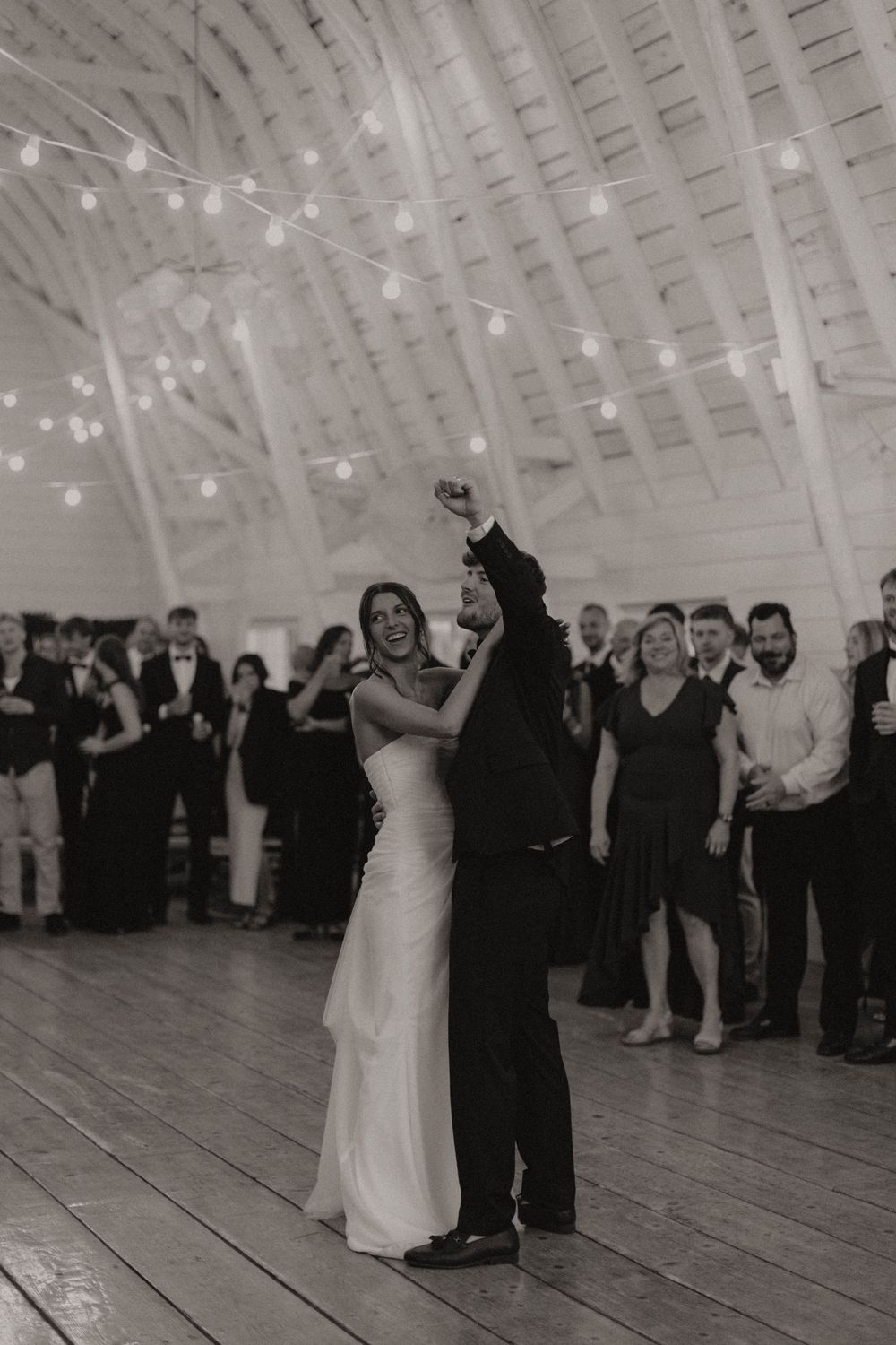 Black and white photo of wedding guests surrounding a dancing couple in a rustic venue.