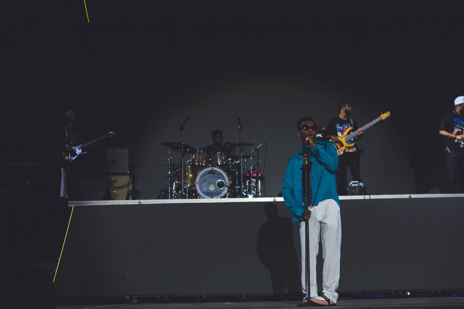A performer in a blue shirt and light pants stands on a dark stage with band members and instruments in the background.