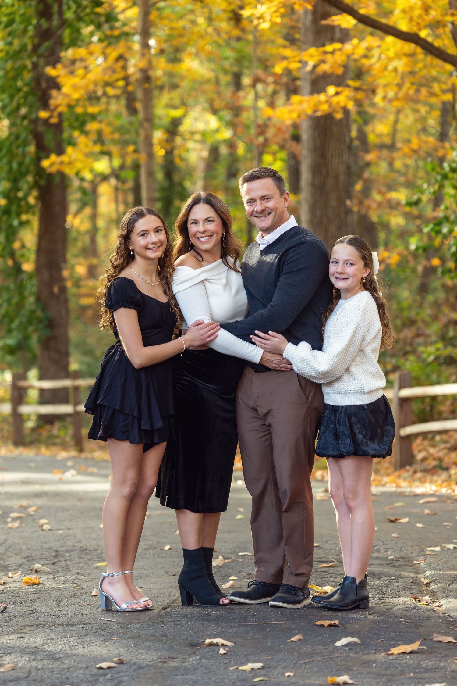 A family embraces while standing outdoors in fall sunshine with a rustic barn in the background.