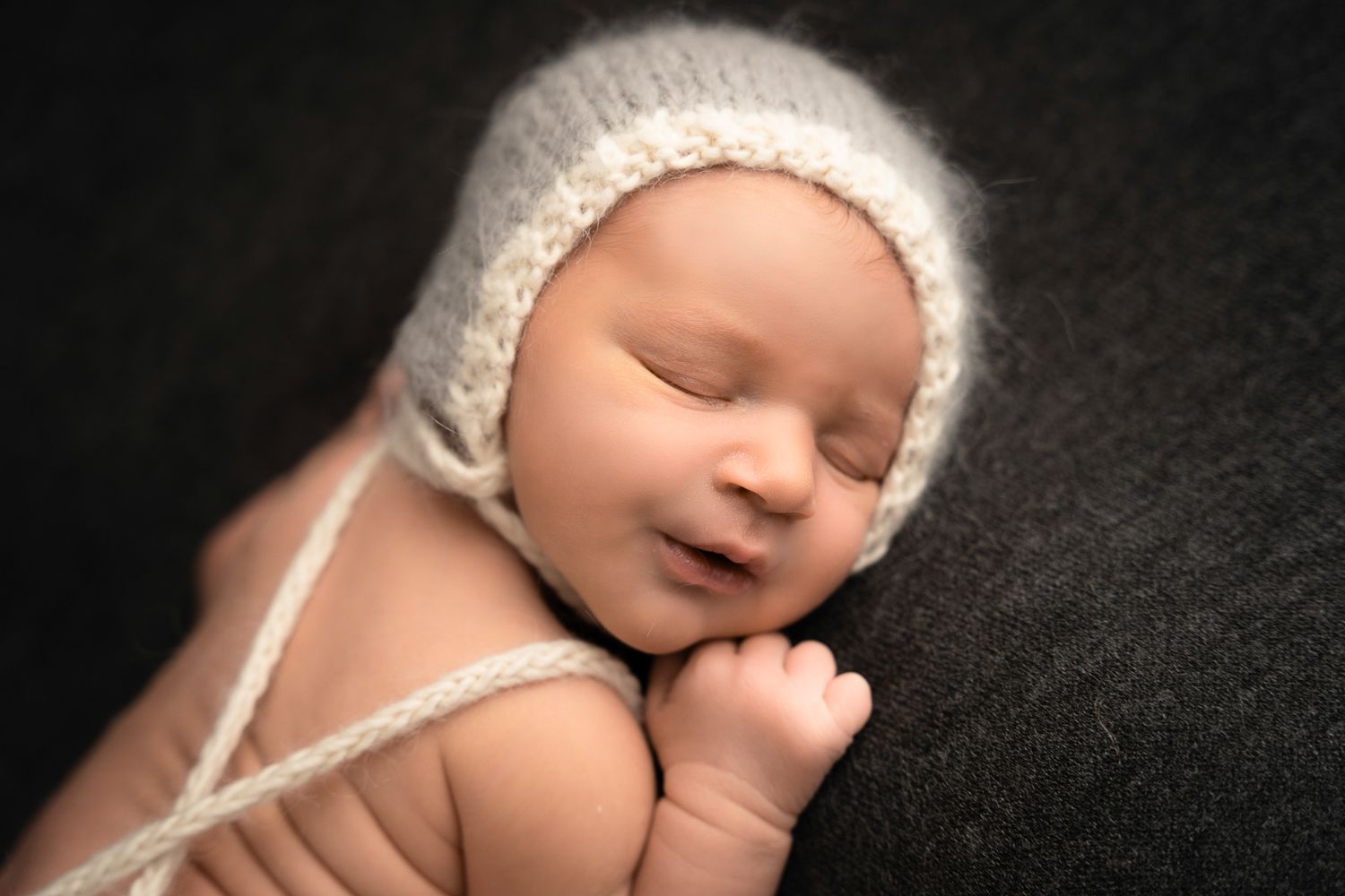 A sleeping newborn wearing a soft white knit bonnet poses against a dark background during posed newborn session in Arlington Heights