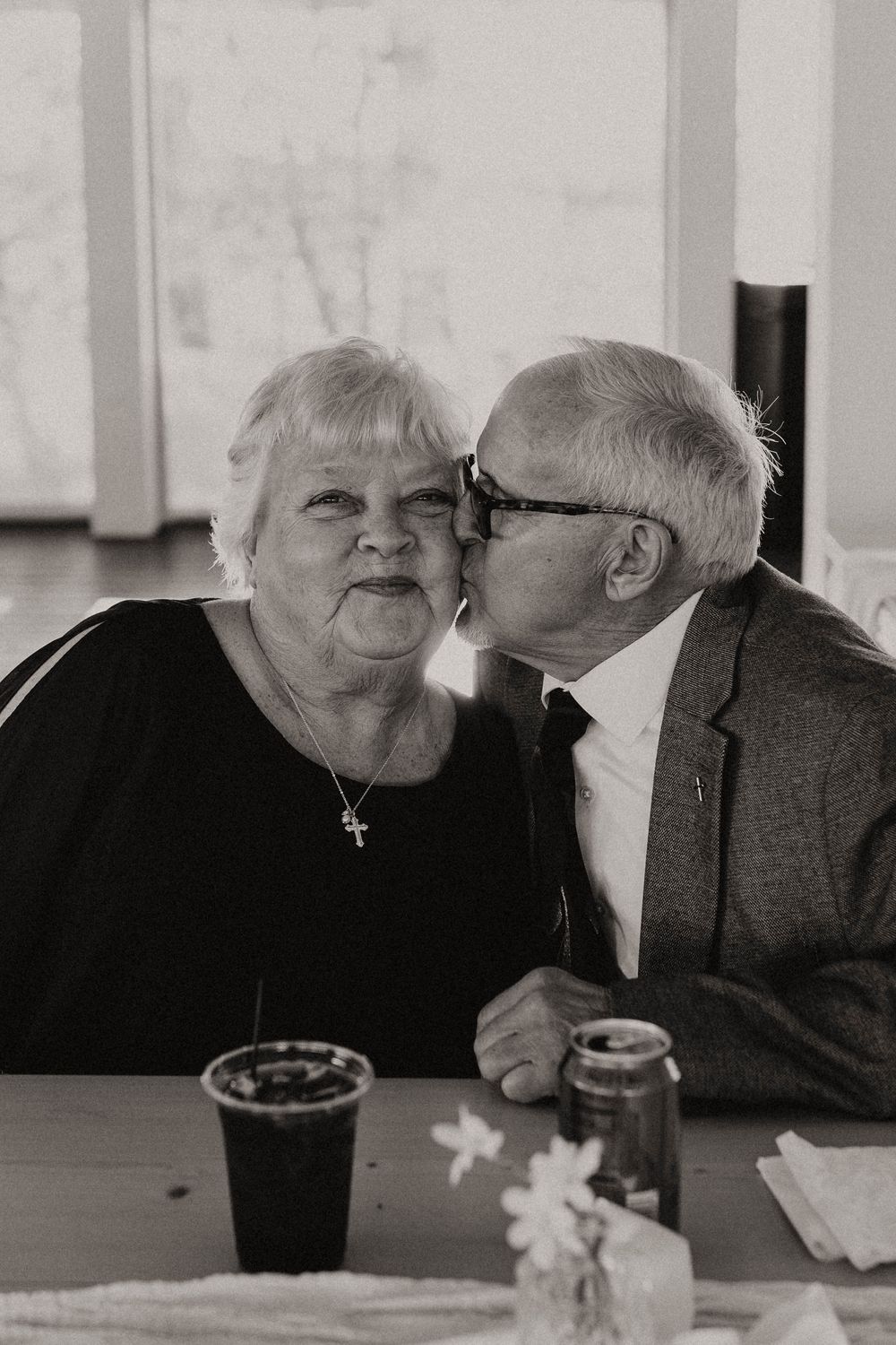 Sweet moment between elderly couple sharing drinks at a table in black and white.