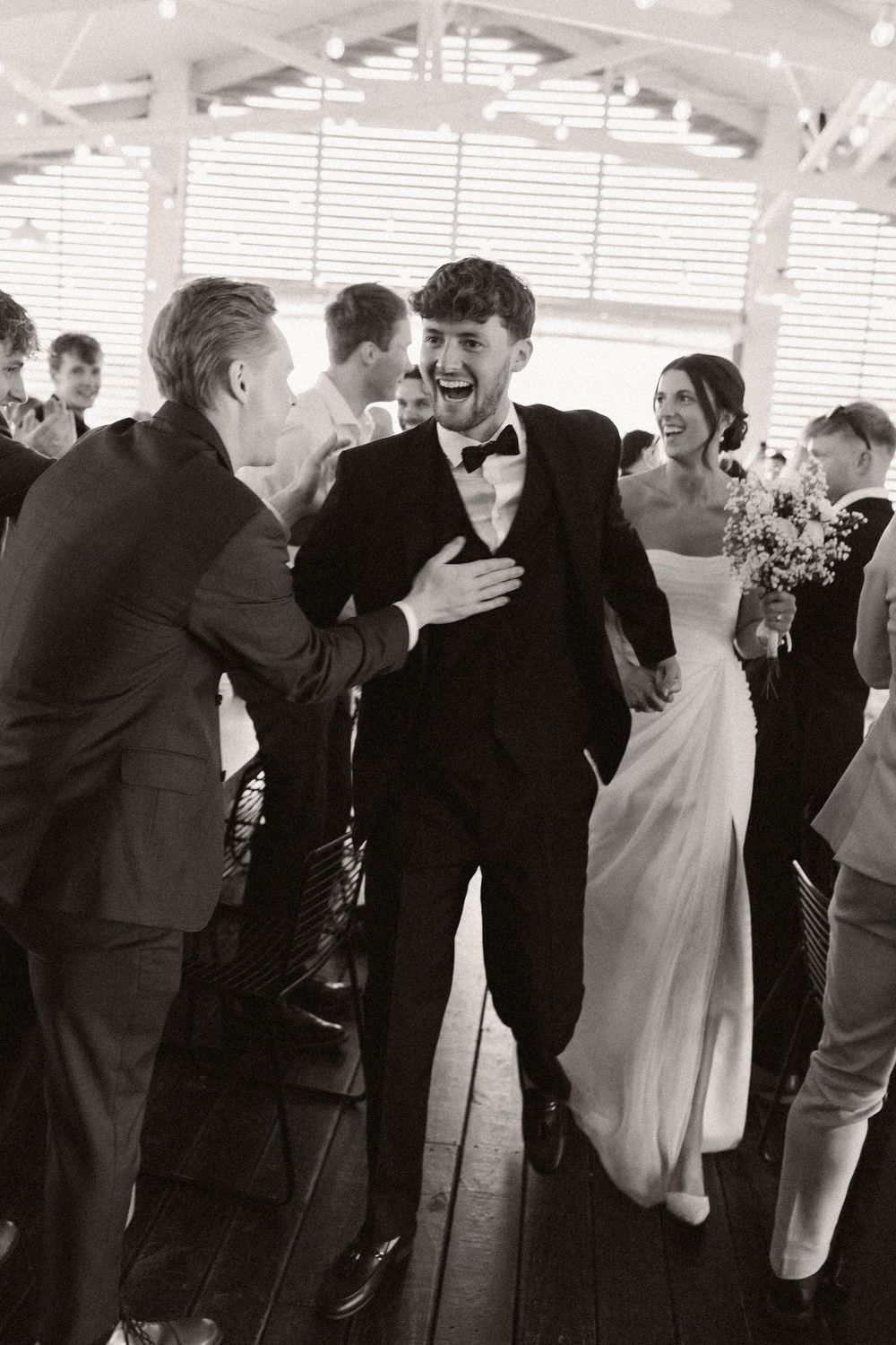 Black and white photo of wedding guests dancing and celebrating in an indoor venue.