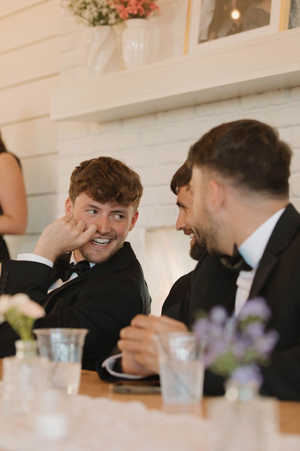 Groomsmen share laughs at an elegant wedding reception with white decor and floral arrangements.