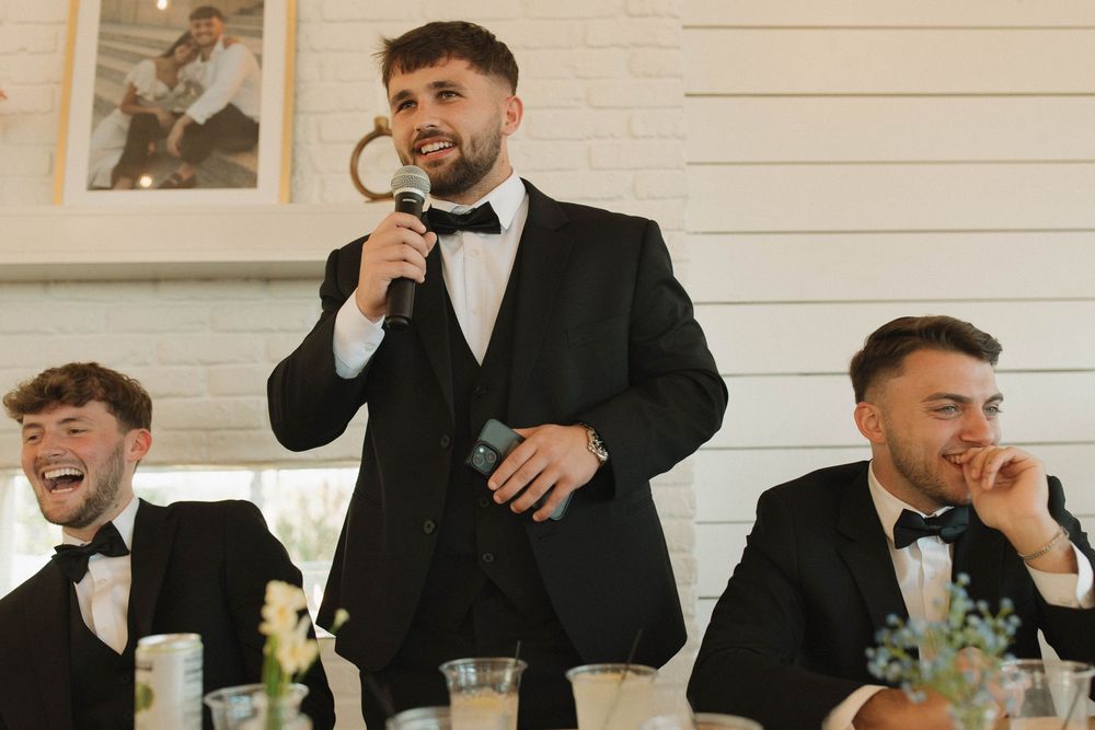 Groomsmen share drinks and laughter at a formal wedding reception with white brick walls.