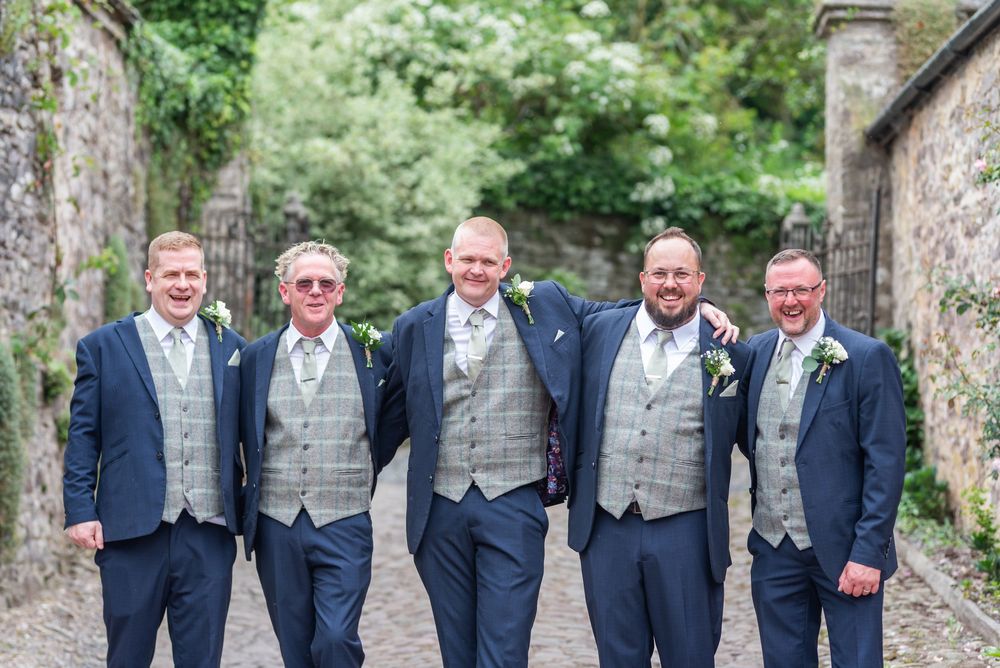 A group of groomsmen wearing navy suits with gray waistcoats pose together outdoors.