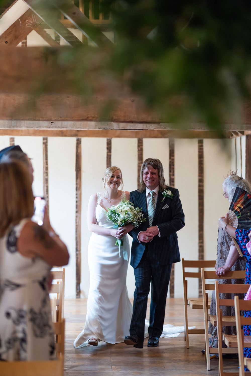 Wedding ceremony taking place in a rustic indoor venue with wooden chairs and beams.