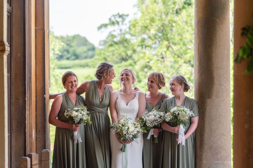 Bridesmaids in sage green dresses stand with bride in colonnade.
