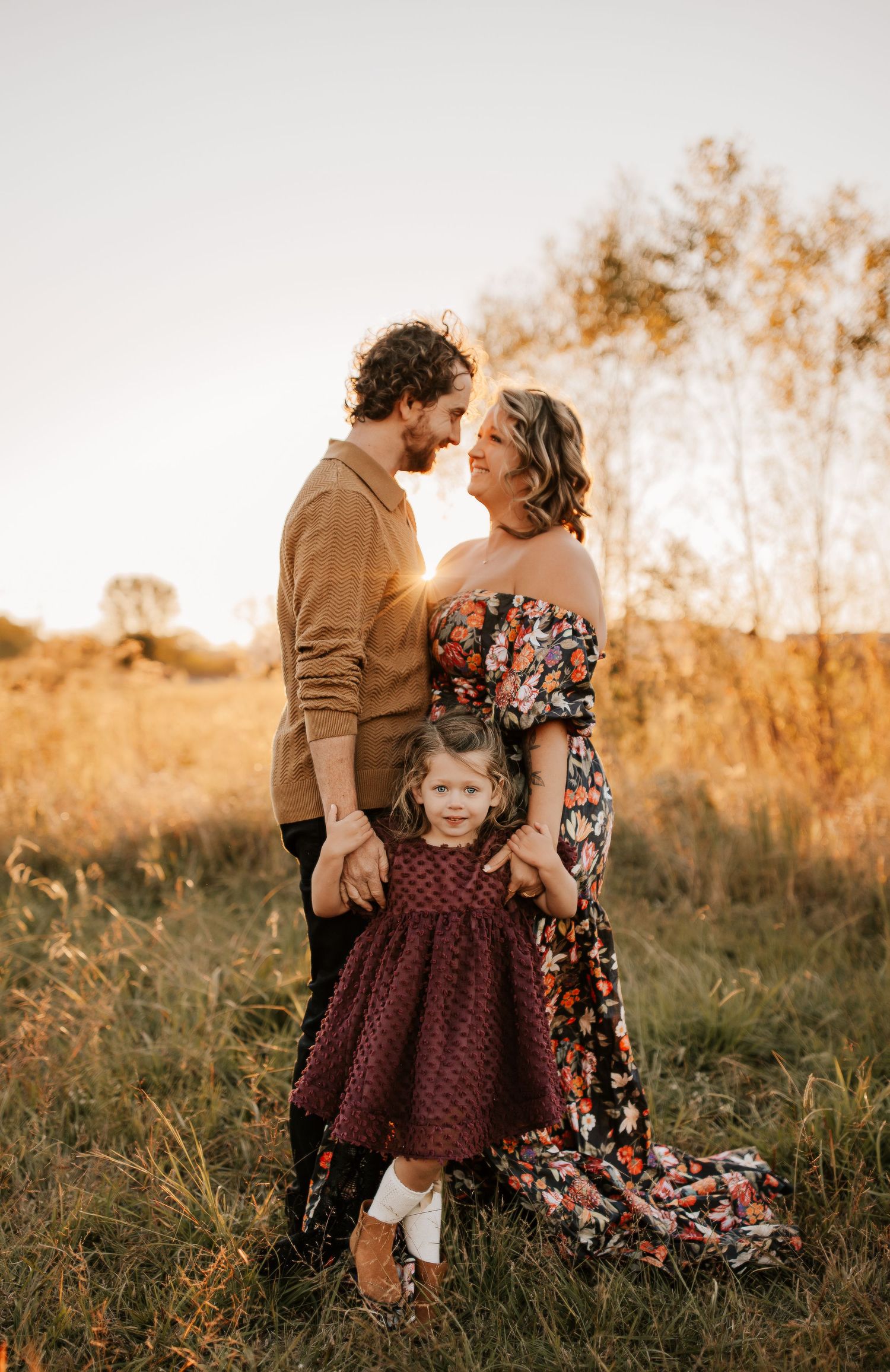 A family wearing brown and floral outfits poses together in a golden sunset-lit field during an outdoor photo session.