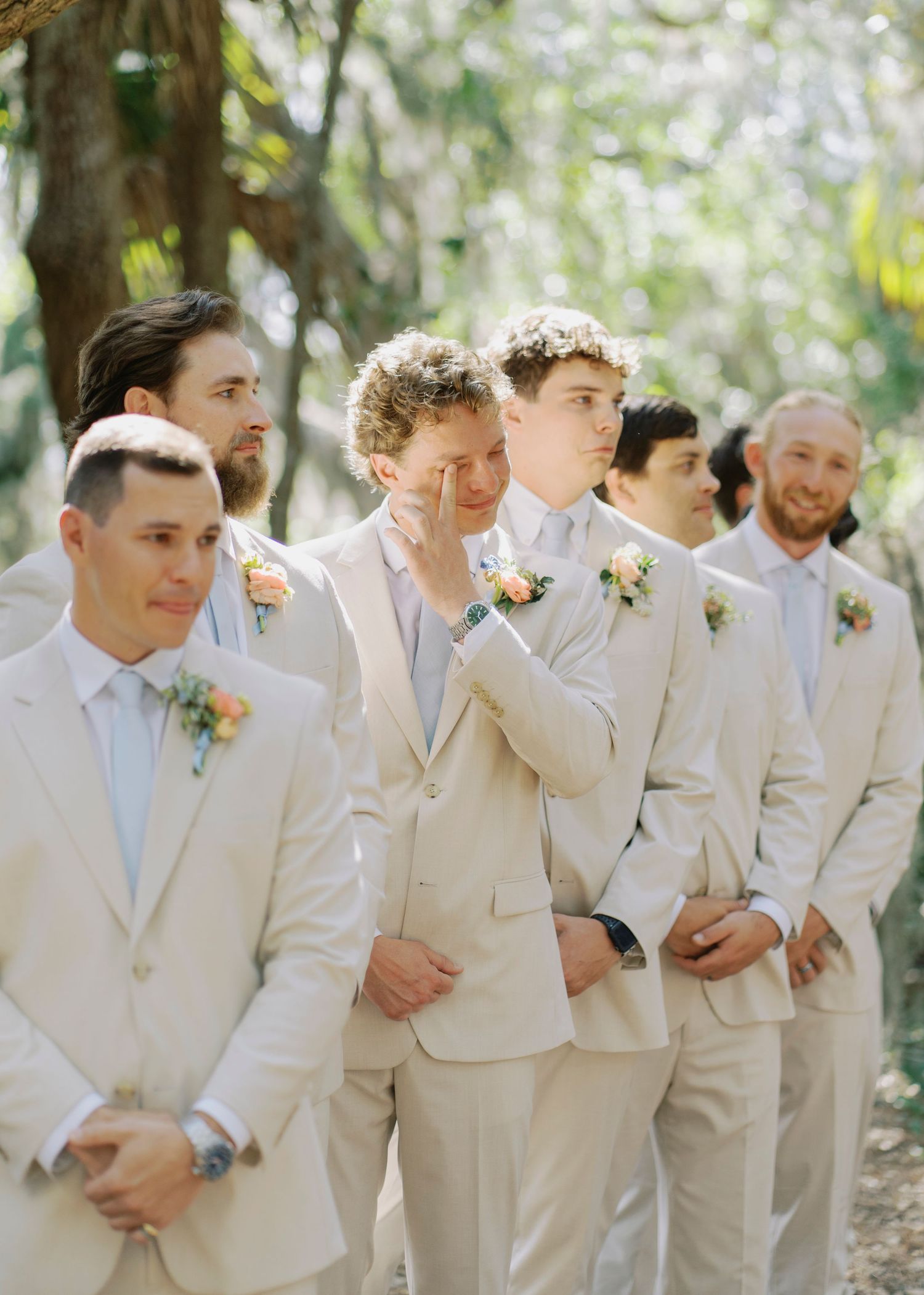 Emotional groomsman wiping tears during outdoor wedding ceremony surrounded by fellow groomsmen.