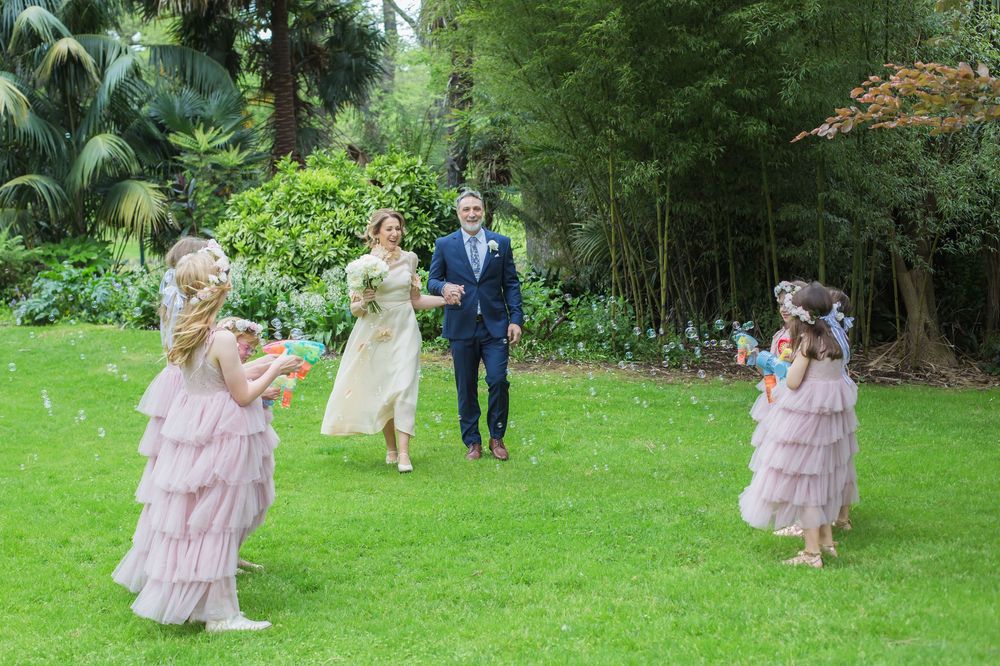 A group of people in formal wear run and play on a lush green lawn surrounded by bamboo and tropical plants.