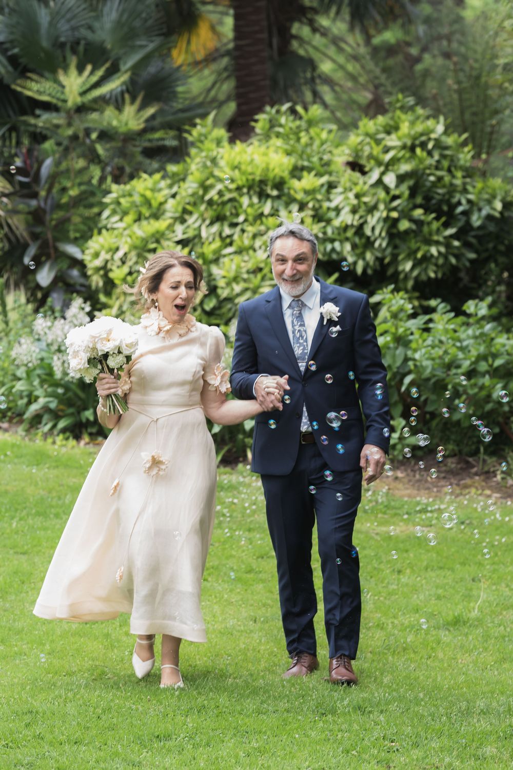 A happy couple walks on a garden lawn while bubbles float around them during their wedding celebration.