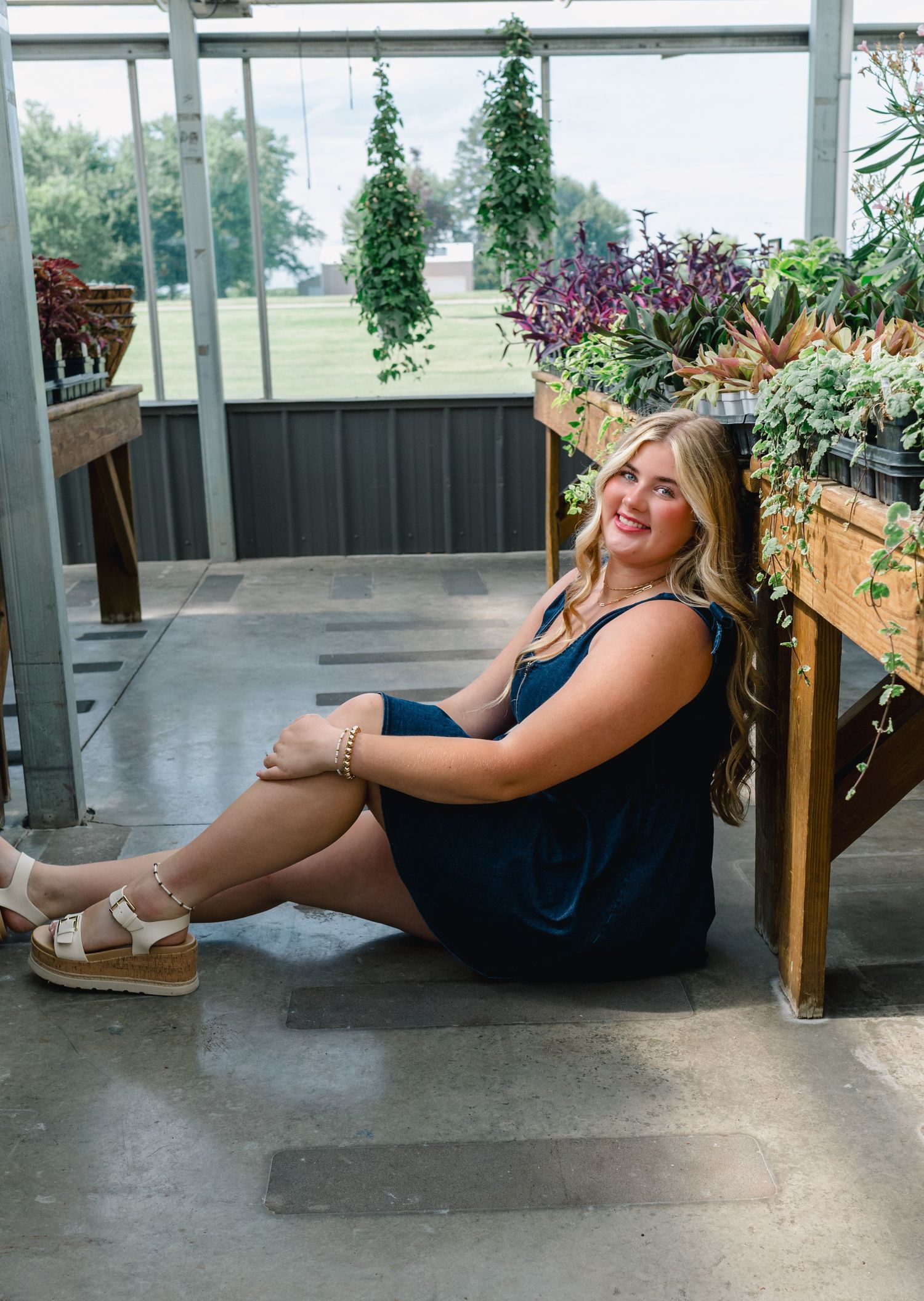 A person in a blue summer dress sits on a covered porch next to decorative planters filled with colorful flowers.