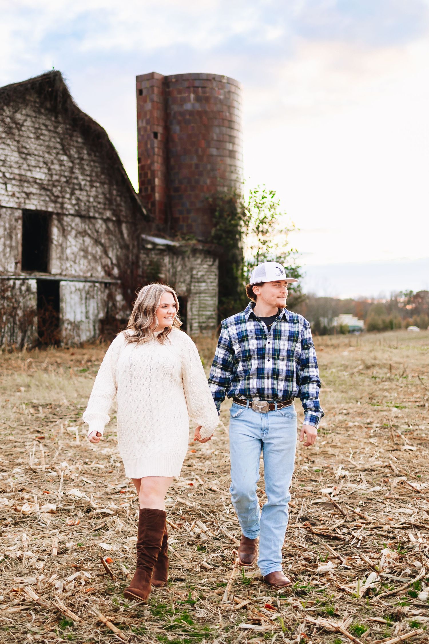 A couple poses in front of old brick farm silos and rustic buildings in a rural field.
