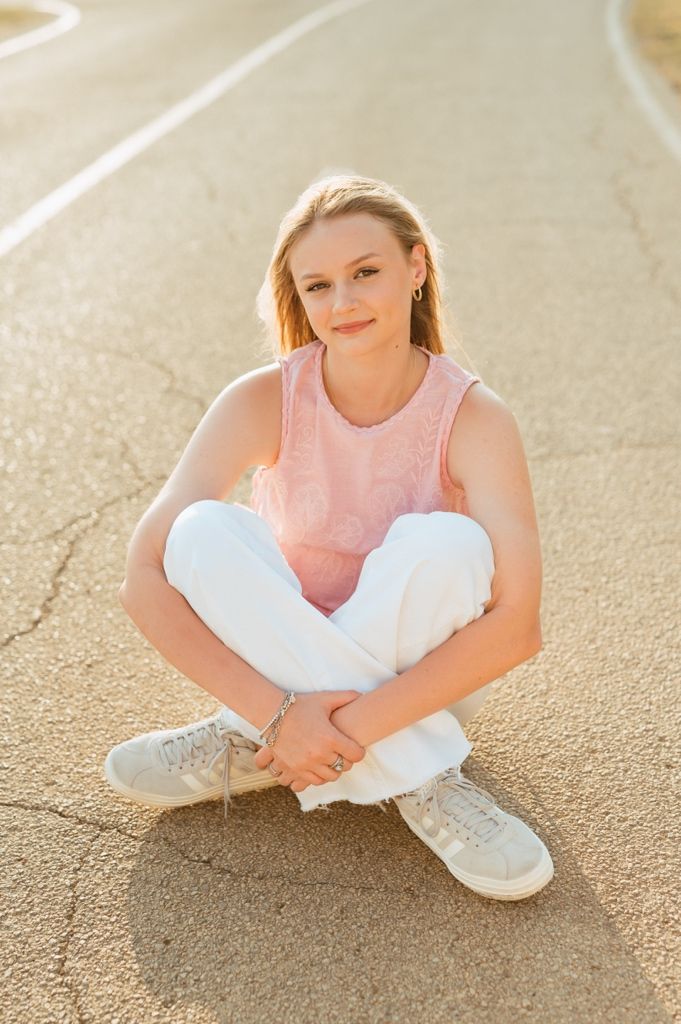 A person in a pink sleeveless top and white pants sits cross-legged on a path during golden hour lighting.