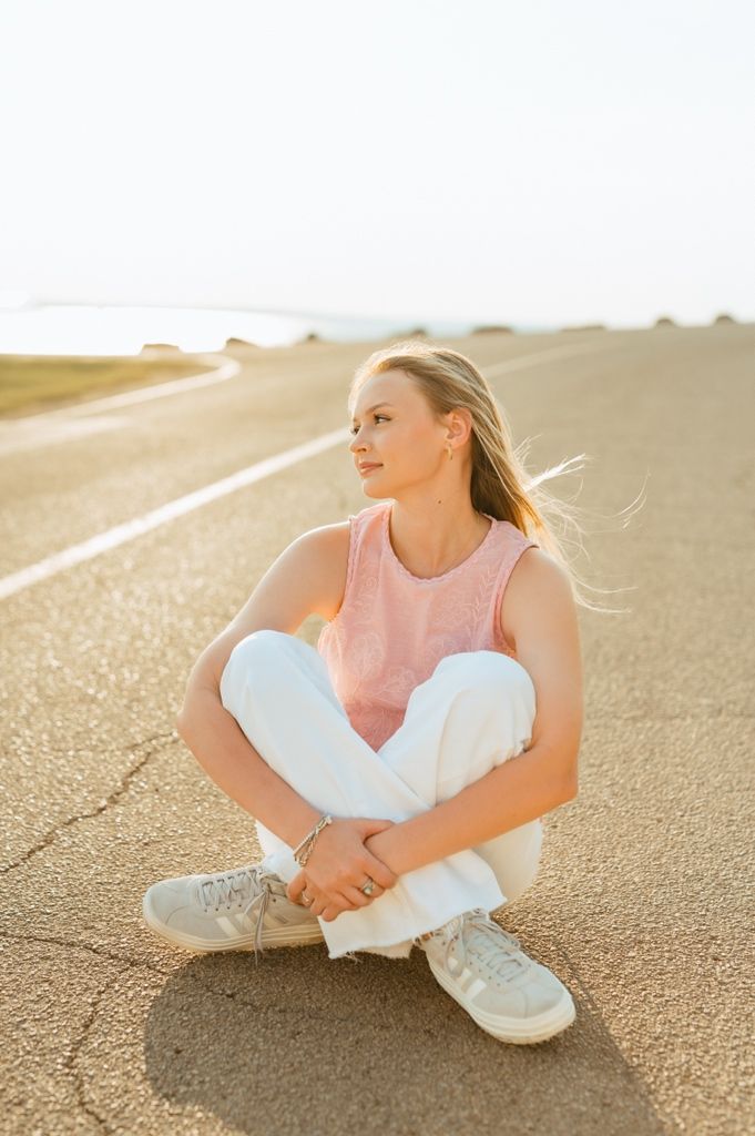 A person in a white outfit sitting on a sunlit beach sand at golden hour with hair blowing in the breeze.