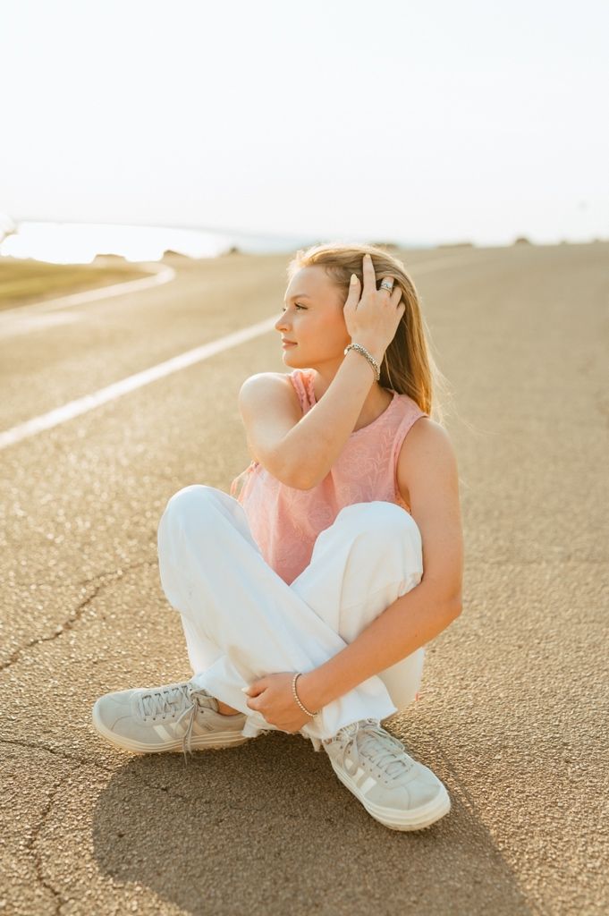 Person in pink sleeveless top and white pants sits on asphalt road during golden hour sunset.