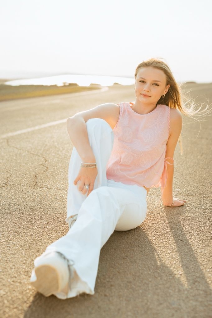 A person in white pants and pink top sits casually on a sandy path during golden hour sunset.