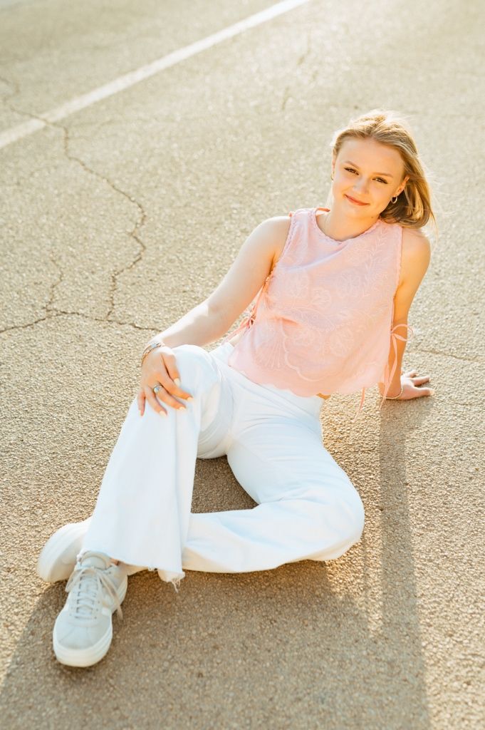 A casual portrait of someone relaxing outdoors wearing a pink sleeveless top, white pants and sneakers.