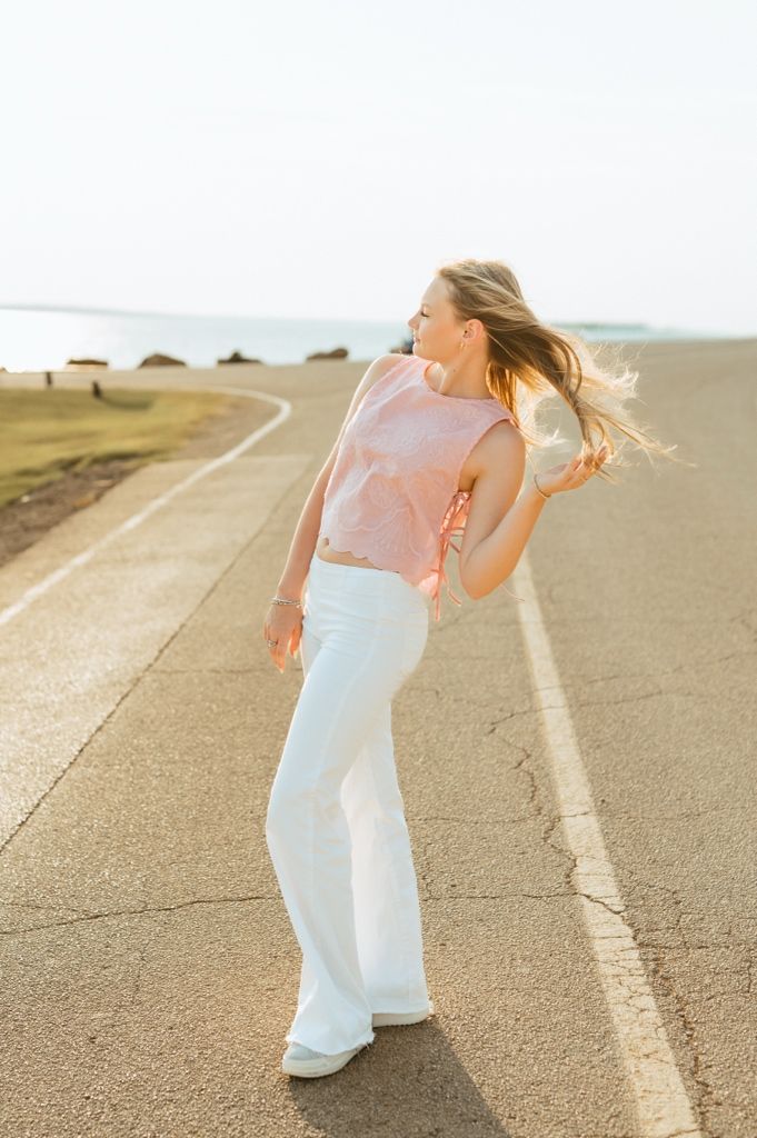 A person in white flared pants and a pink top stands on a road during golden hour while their hair blows in the wind.