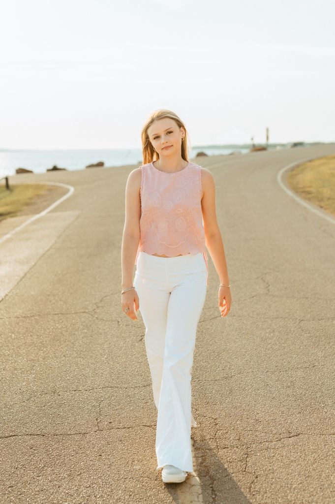 A person wearing a pink sleeveless top and white pants walks down a curved road at sunset.