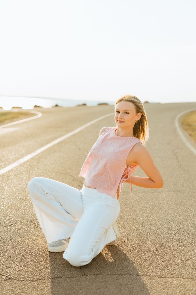 A person in a pink sleeveless top and white pants kneels on a curving desert road at sunset.