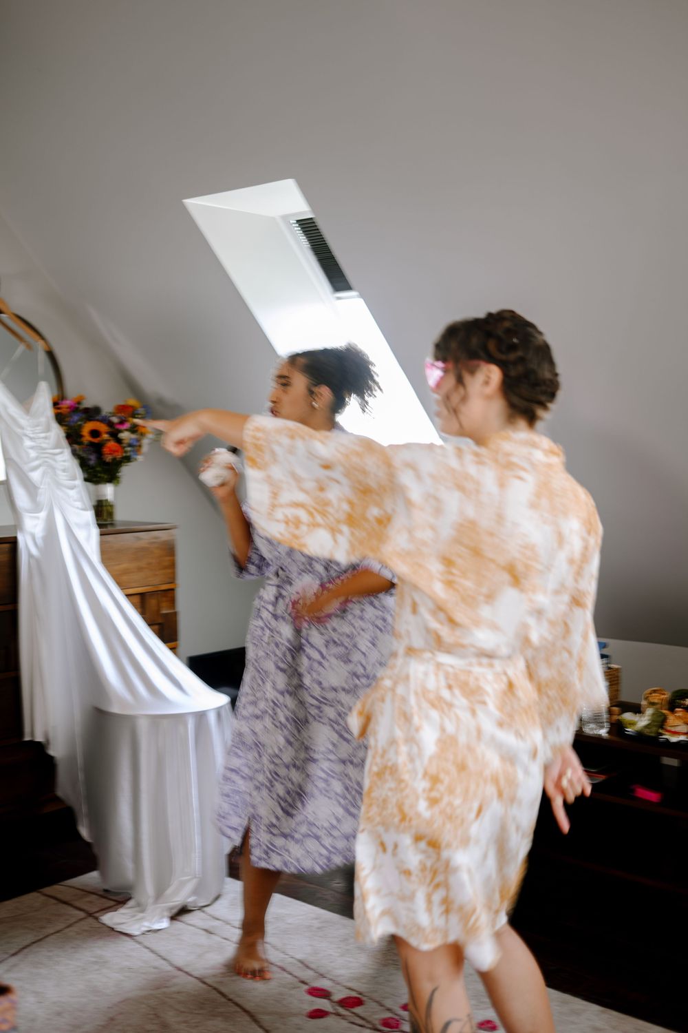 Two women in robes dance together in a bright room decorated with flowers.