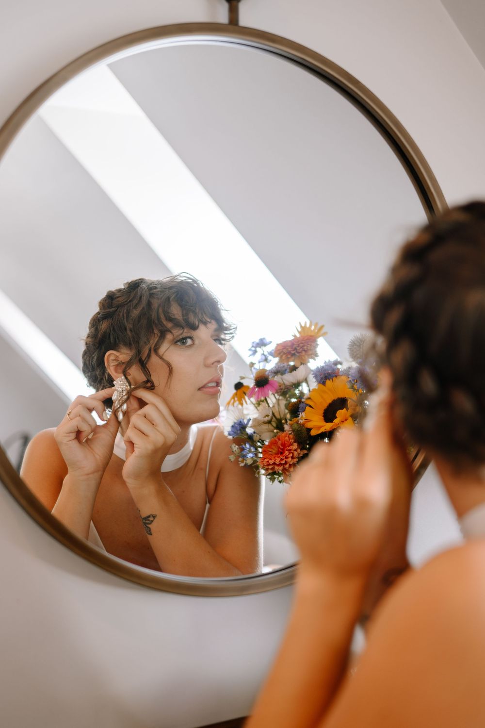 Someone putting on earrings while looking in a round mirror with a colorful floral arrangement visible.