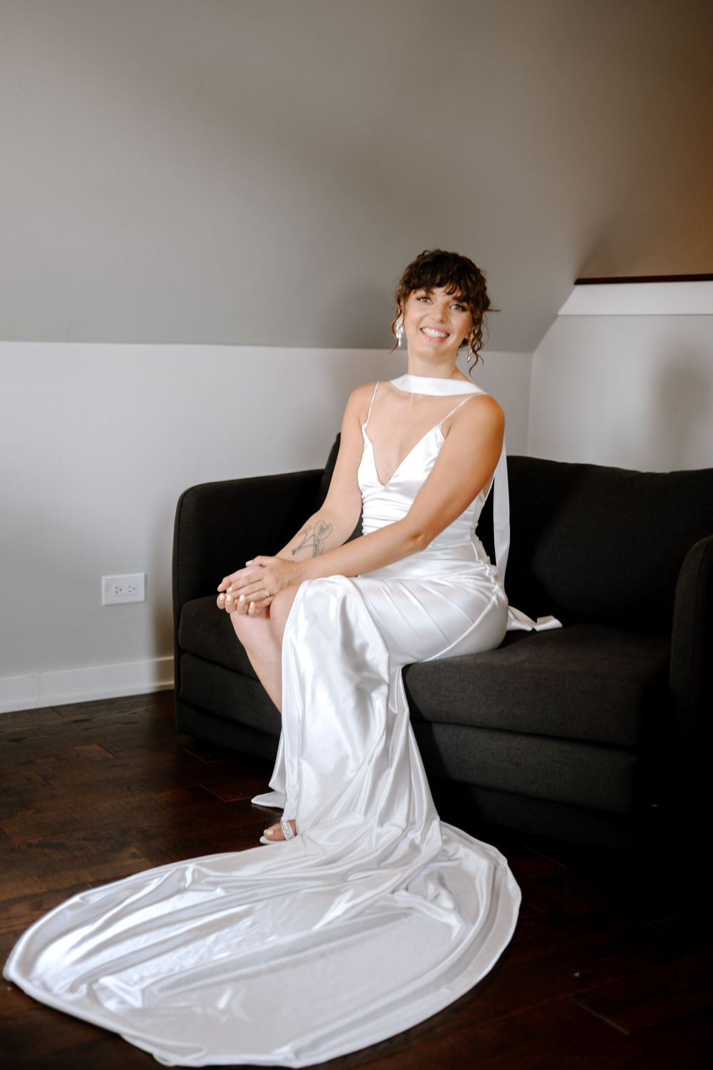 Figure in flowing white dress sits gracefully on dark sofa with dramatic lighting in contemporary interior setting.