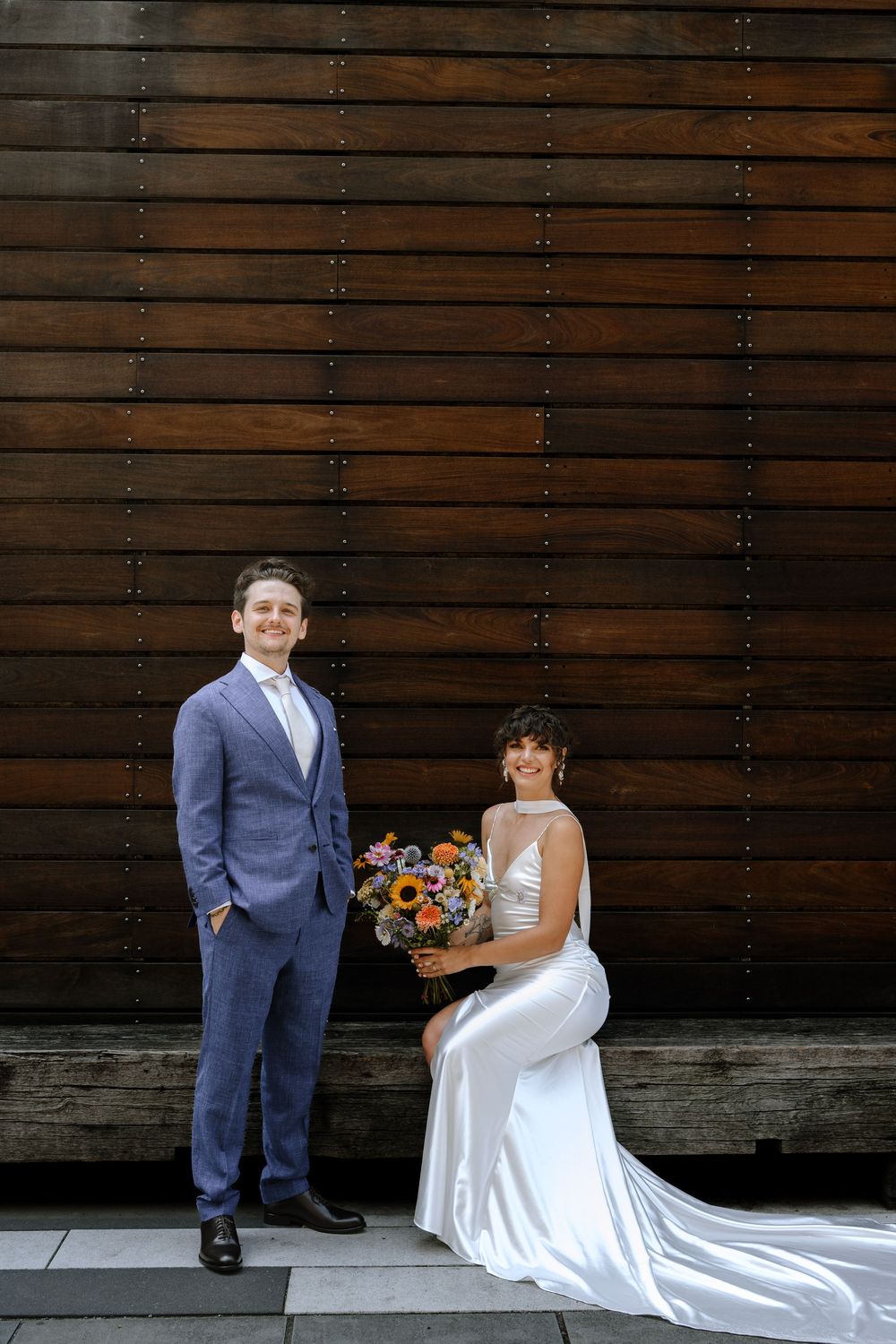 Newlywed couple poses against dark wooden wall with long white wedding dress train and navy suit.
