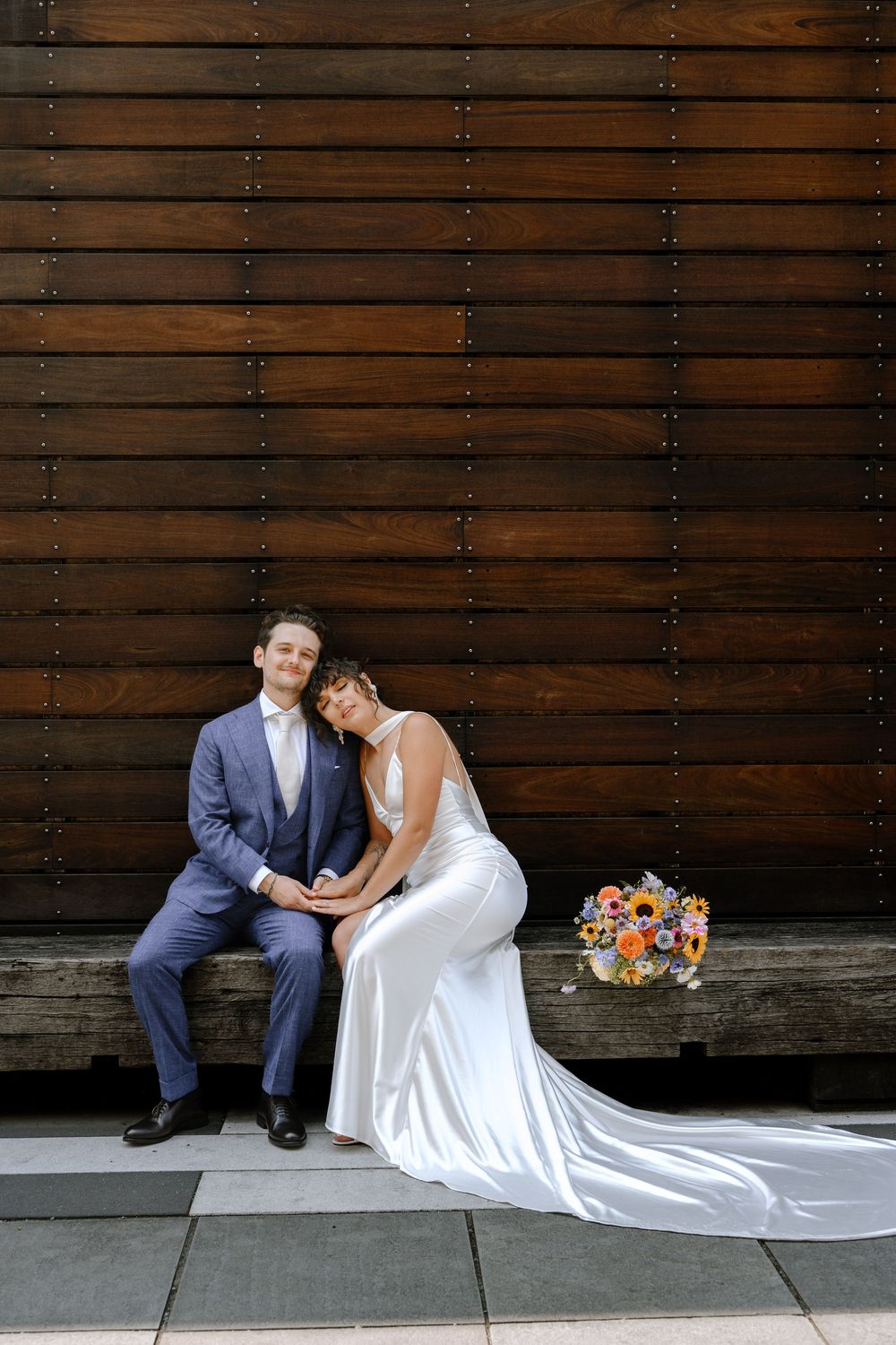 Romantic wedding couple sitting on concrete with flowing white dress and colorful bouquet against wooden wall.
