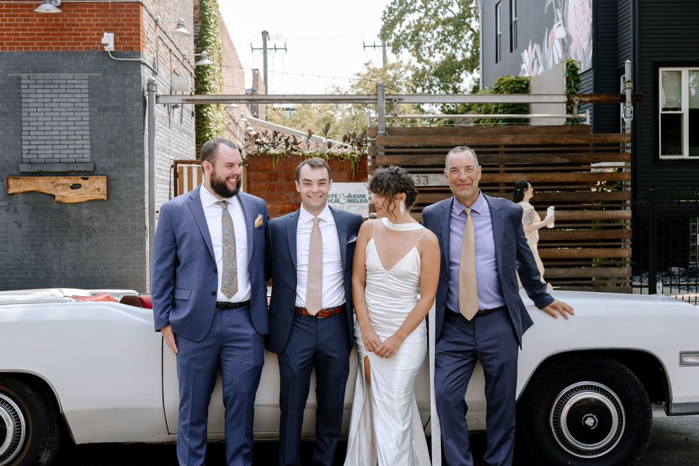 A wedding party in formal attire stands in front of a vintage white car outside a modern industrial building.