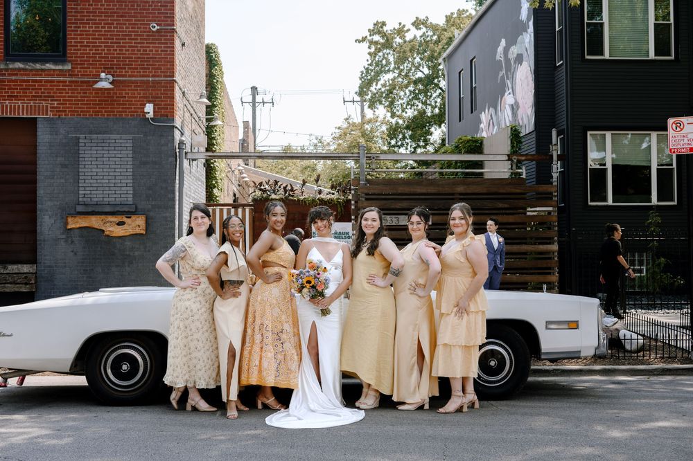 Wedding party in champagne colored dresses poses together outside brick building on sunny day.