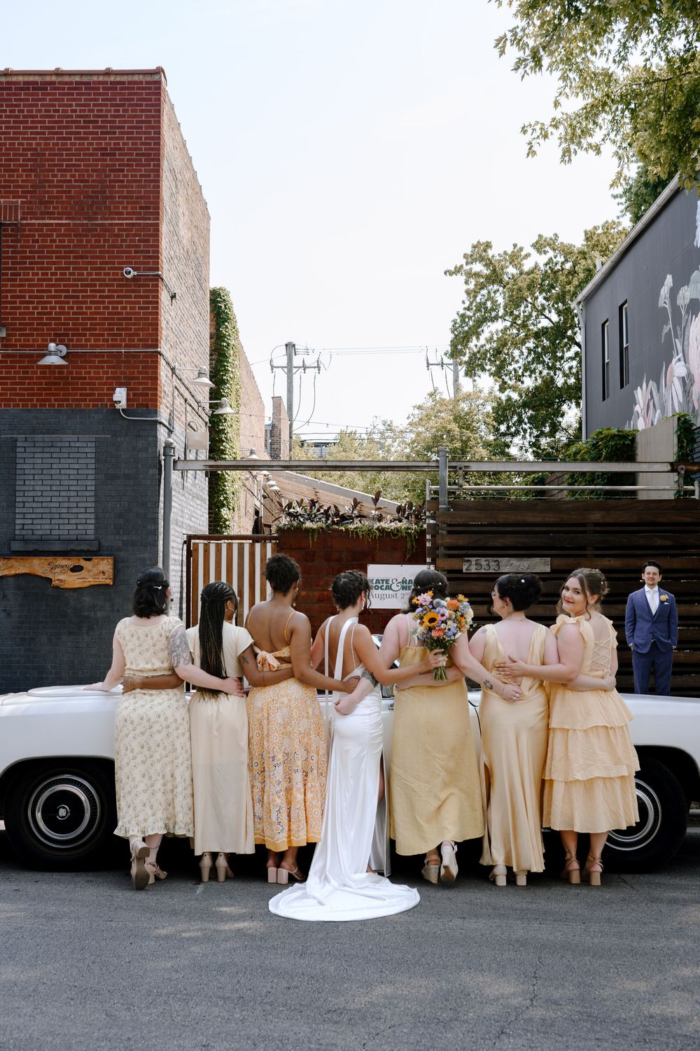 Wedding party in champagne-colored dresses posing in line against brick wall backdrop.
