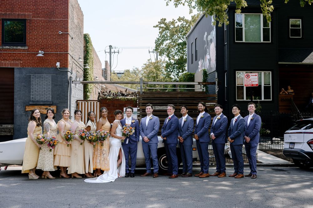 A wedding party in formal attire poses outdoors in an urban setting with brick buildings and trees in the background.