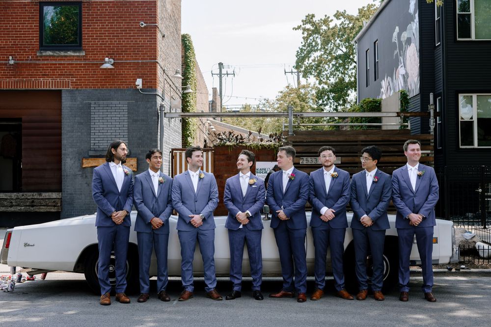 Groomsmen in navy suits gather for formal wedding photos against various outdoor backdrops and brick buildings.