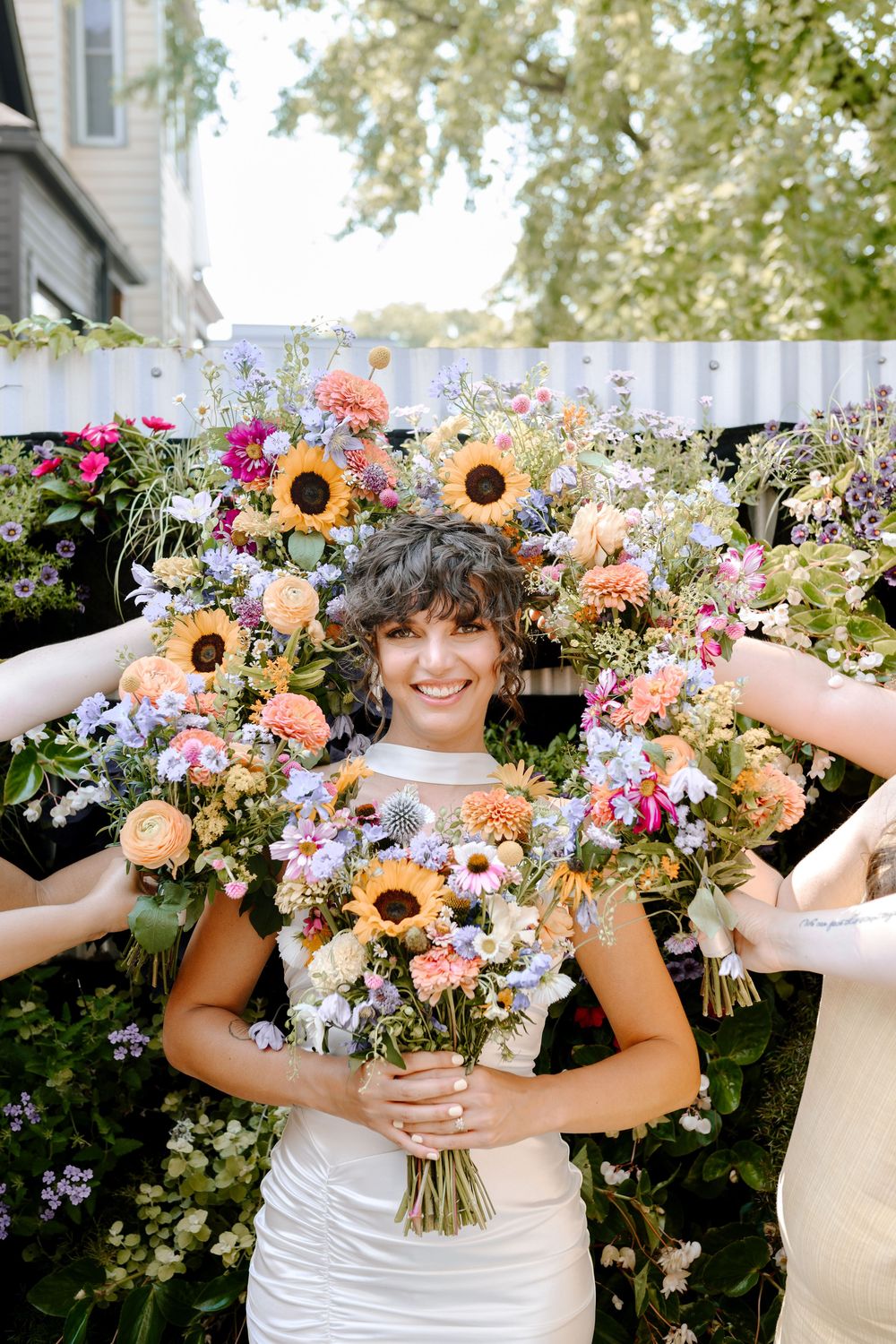 Wedding party holds vibrant wildflower bouquets with sunflowers against white building backdrop.