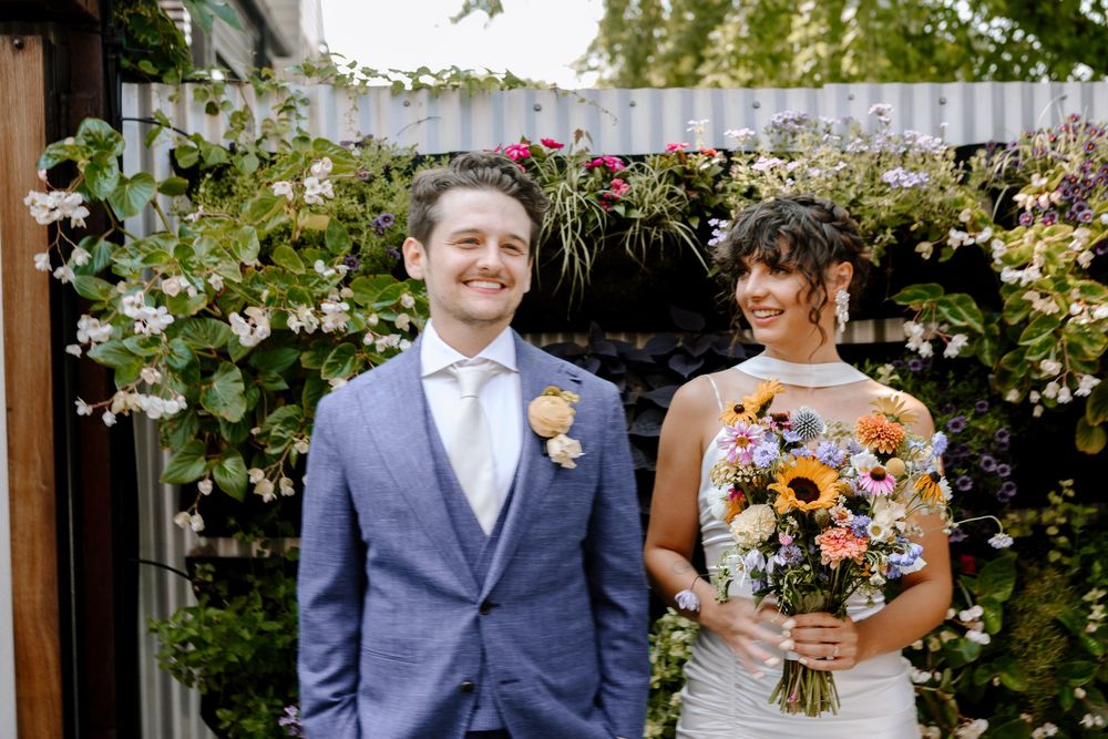 A wedding couple stands in a garden setting with floral decorations and greenery in the background.