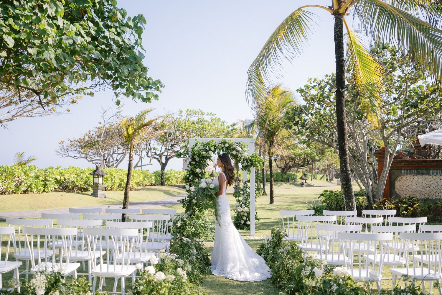 White wedding chairs arranged outdoors under palm trees for a tropical beach ceremony with ocean views in the background.