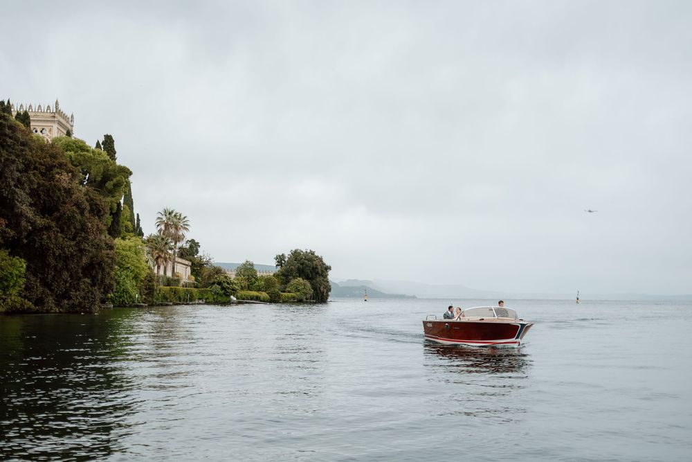 A wooden boat cruises past a historic castle perched on the misty shores of Lake Garda Italy. Destination wedding photographer.
