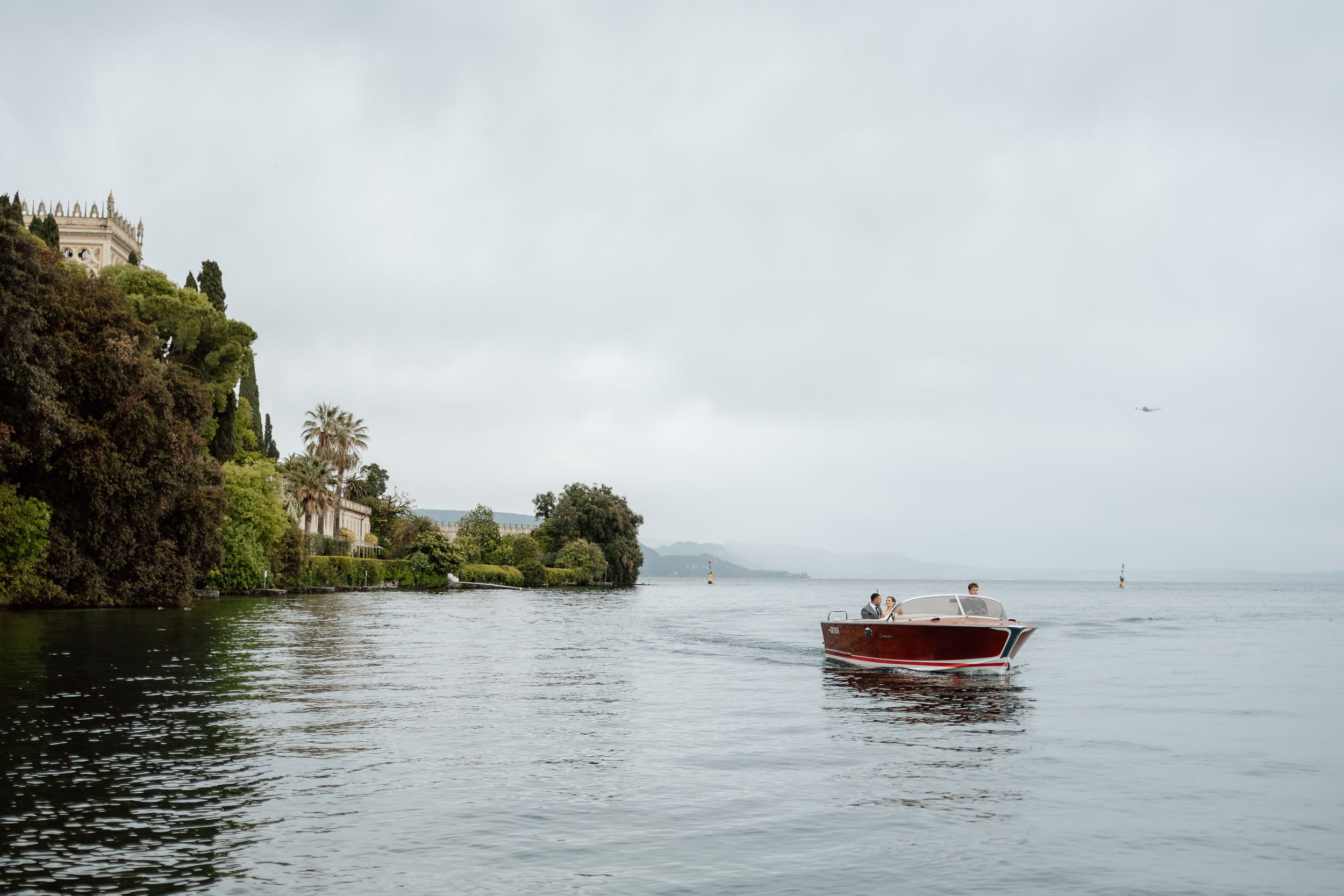 A wooden boat cruises past a historic castle perched on the misty shores of Lake Garda Italy. Destination wedding photographer.