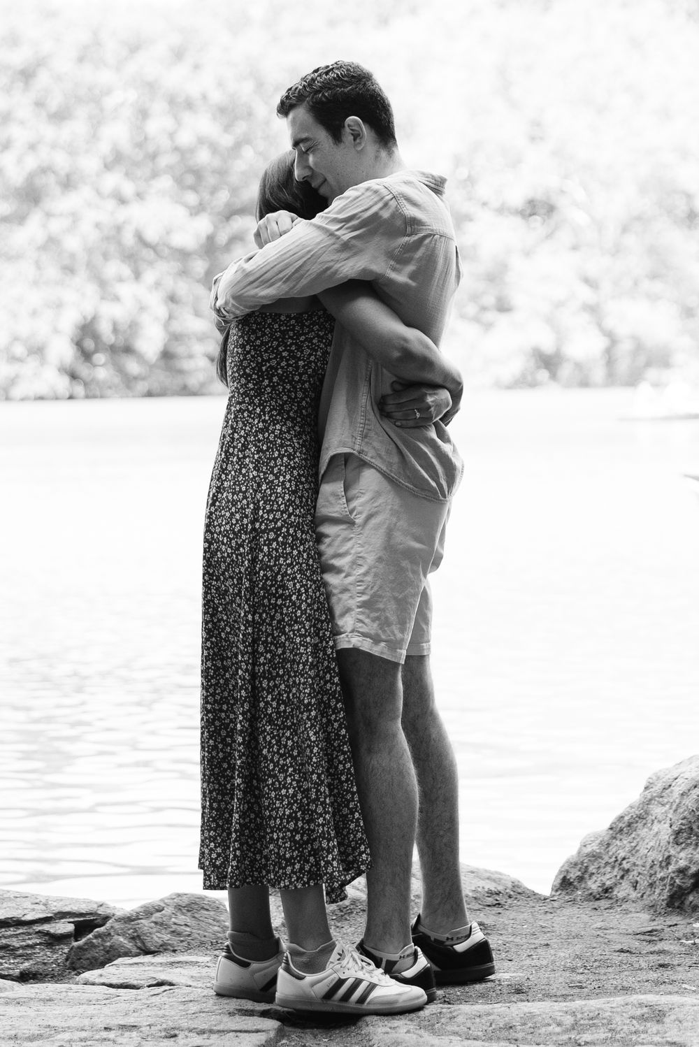 A romantic black and white photo of a couple embracing by the water's edge after a NYC surprise proposal in Central Park.
