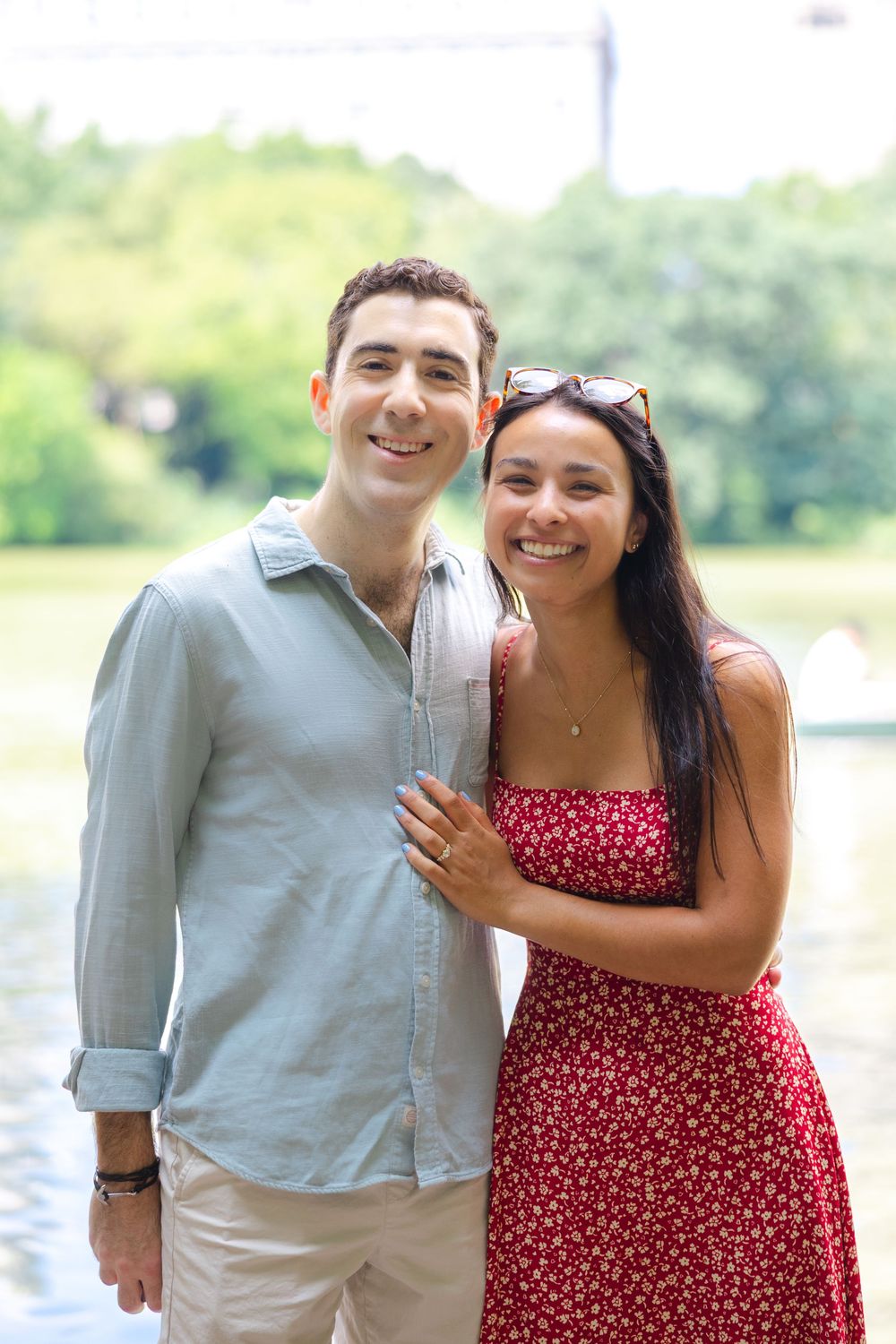 An engaged couple poses together after their proposal with the woman wearing a red dress and sunglasses and the man wearing a collared shirt