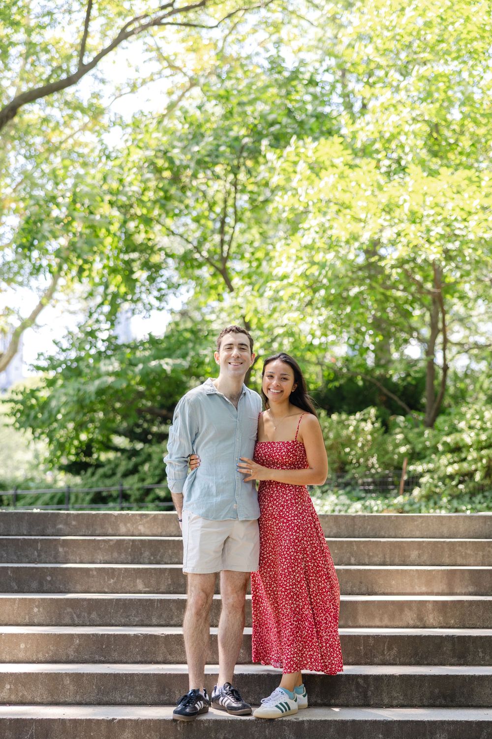A couple stands together on outdoor stone steps in Central Park surrounded by lush green trees on a sunny summer day.