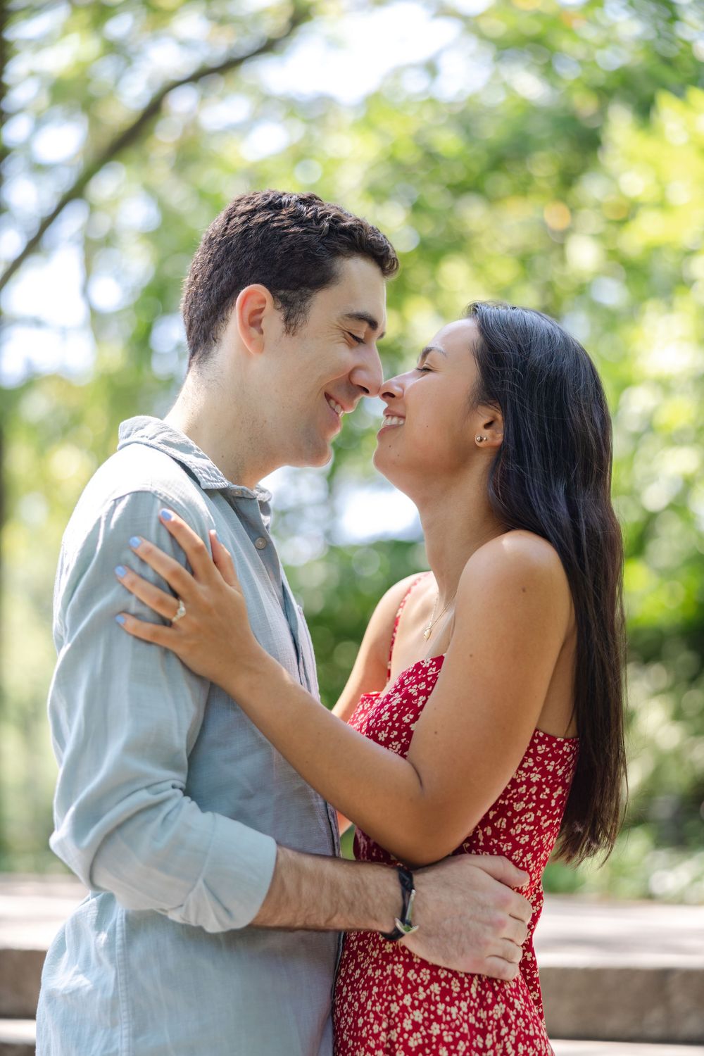 A couple shares an intimate moment in Central Park, with her wearing a red floral dress and him in a light blue shirt.