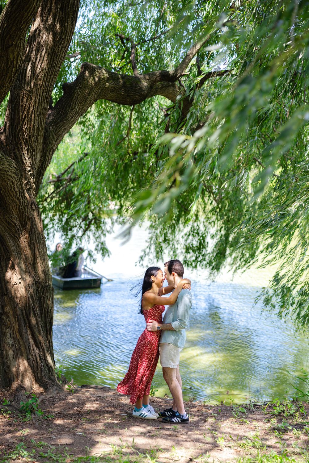 A newly engaged couple kisses beneath a willow tree by the tranquil Central Park Lake on a summer afternoon after their proposal.