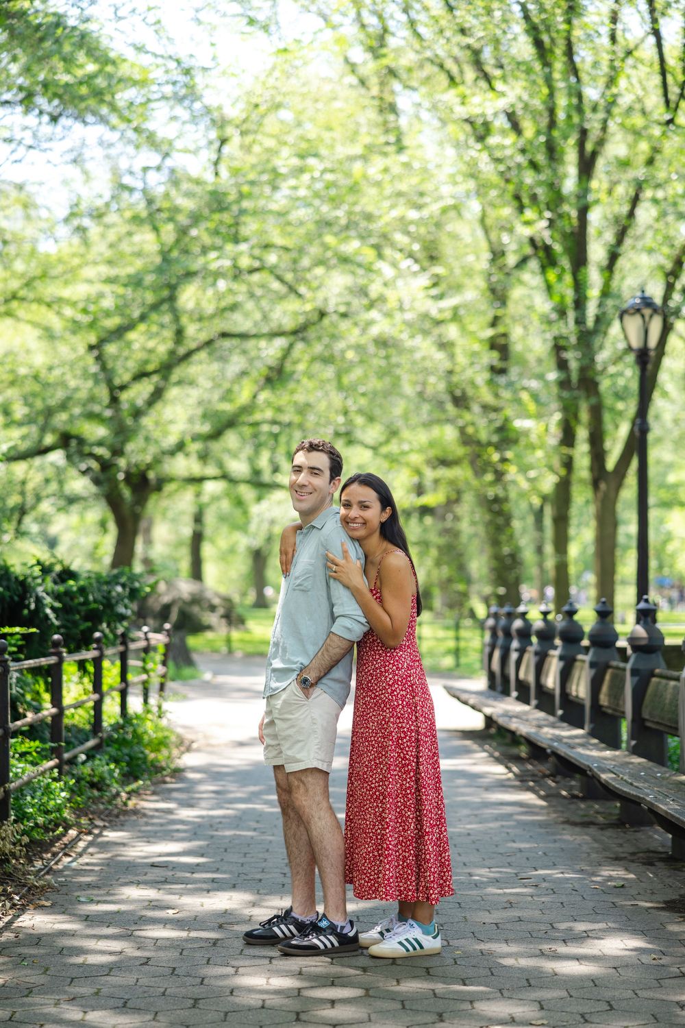 A couple shares a romantic moment on a tree-lined pathway in the Central Park Mall area on a sunny summer day after their surprise proposal.