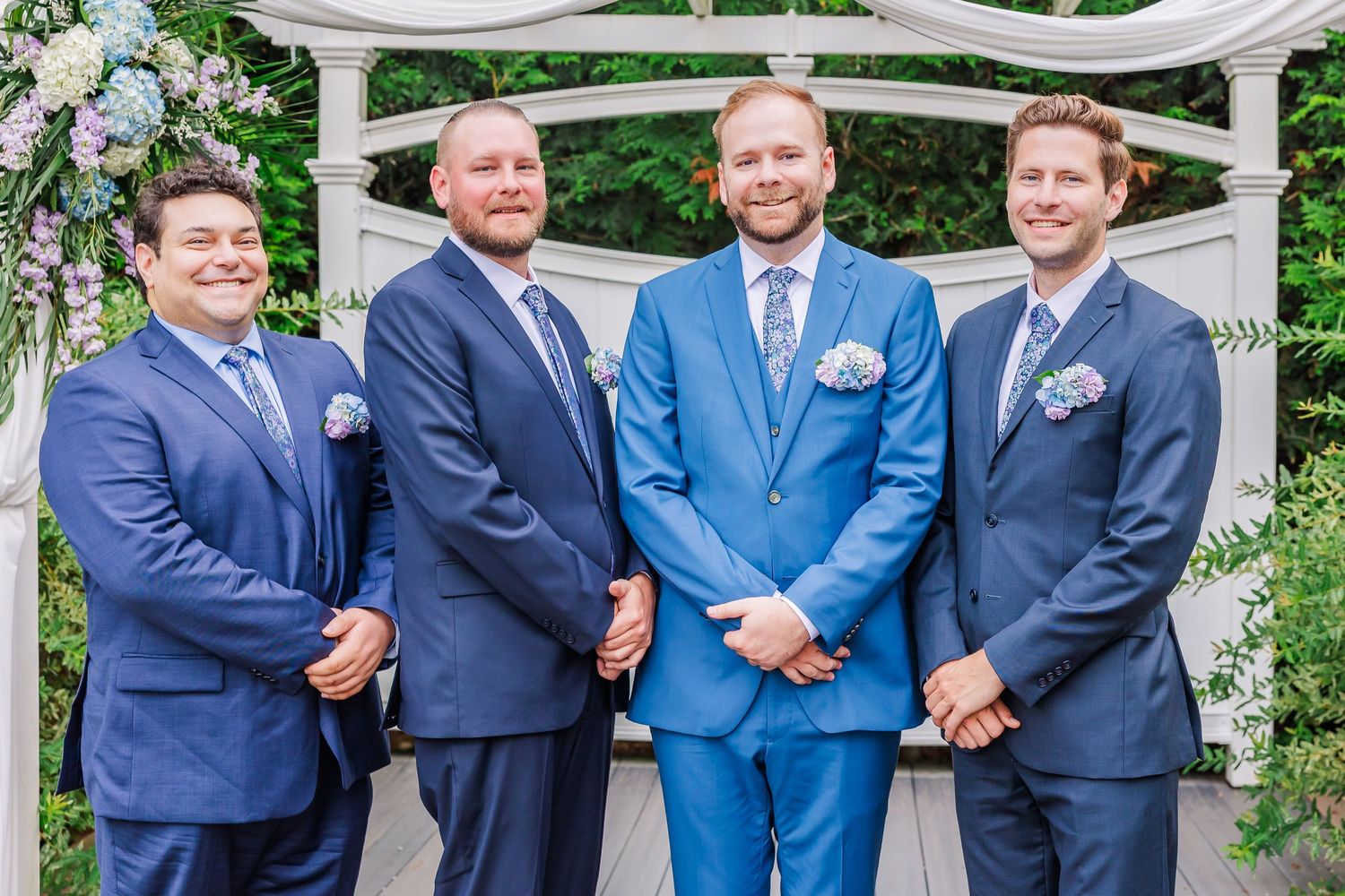 Groomsmen in coordinating navy and bright blue suits stand together under a white arbor at an outdoor wedding ceremony.