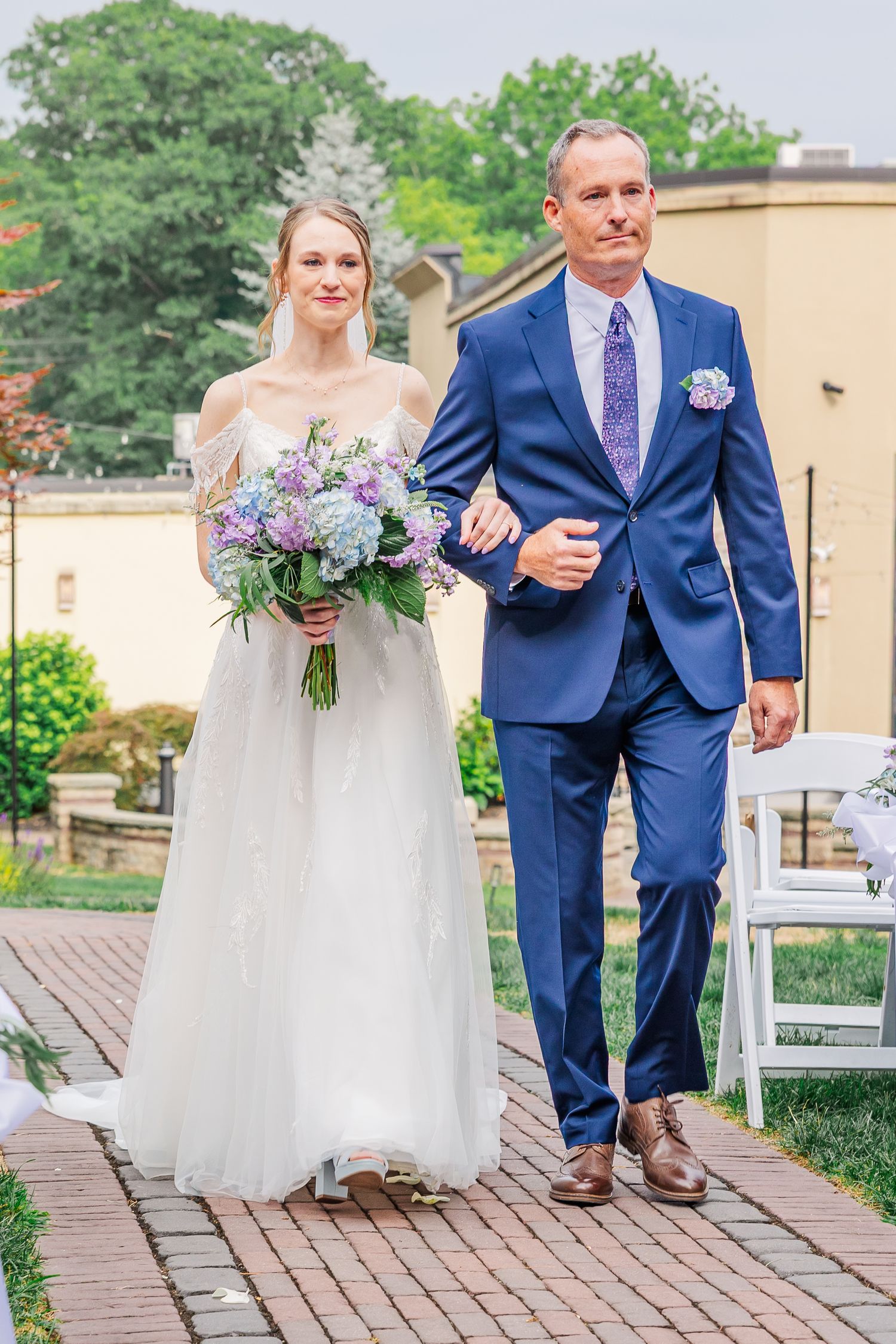 Couple walking along wooden boardwalk in wedding attire with blue suit and white dress at an outdoor venue.