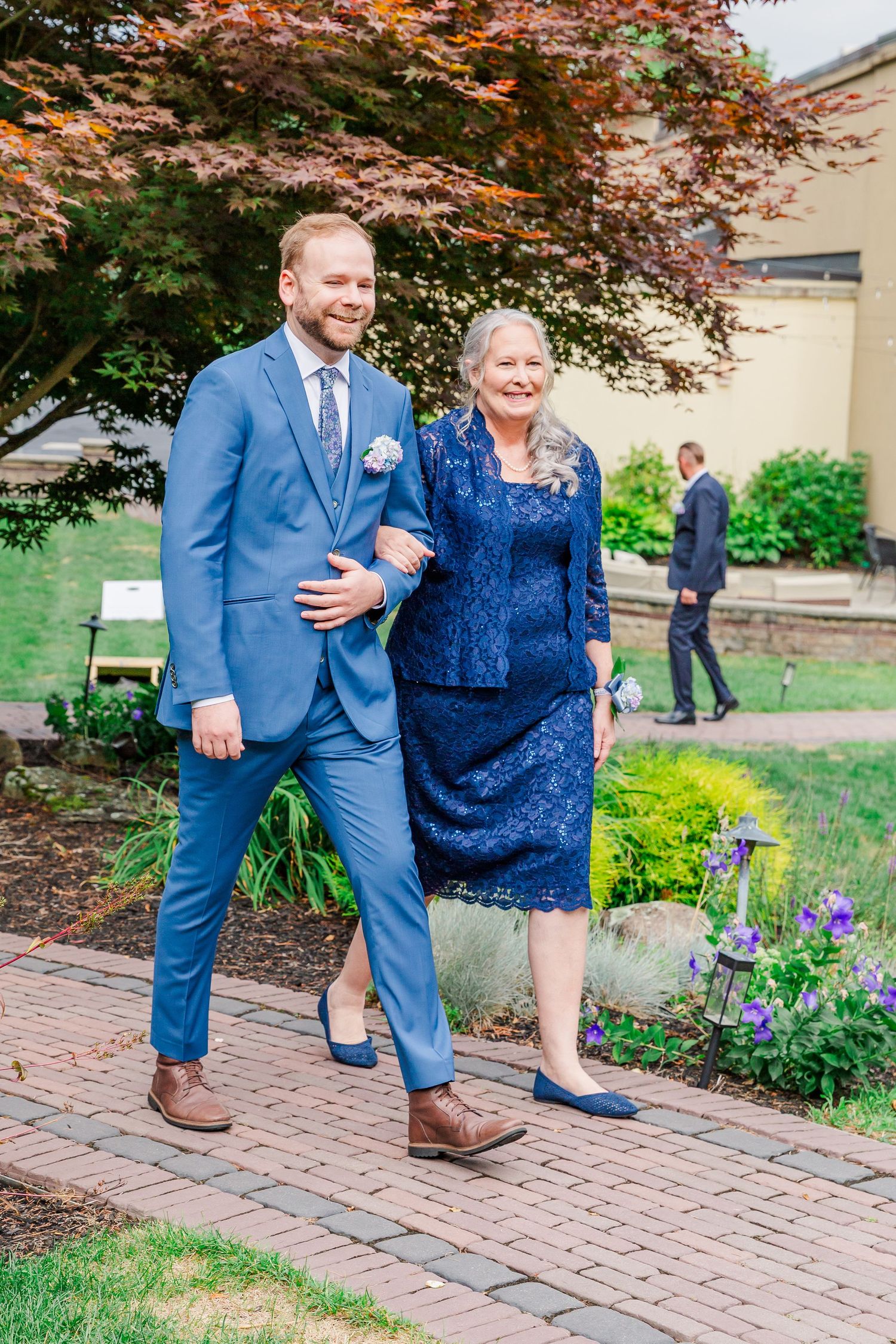 A formal wedding moment walking along a brick path under a Japanese maple tree with blooming flowers at an outdoor venue.