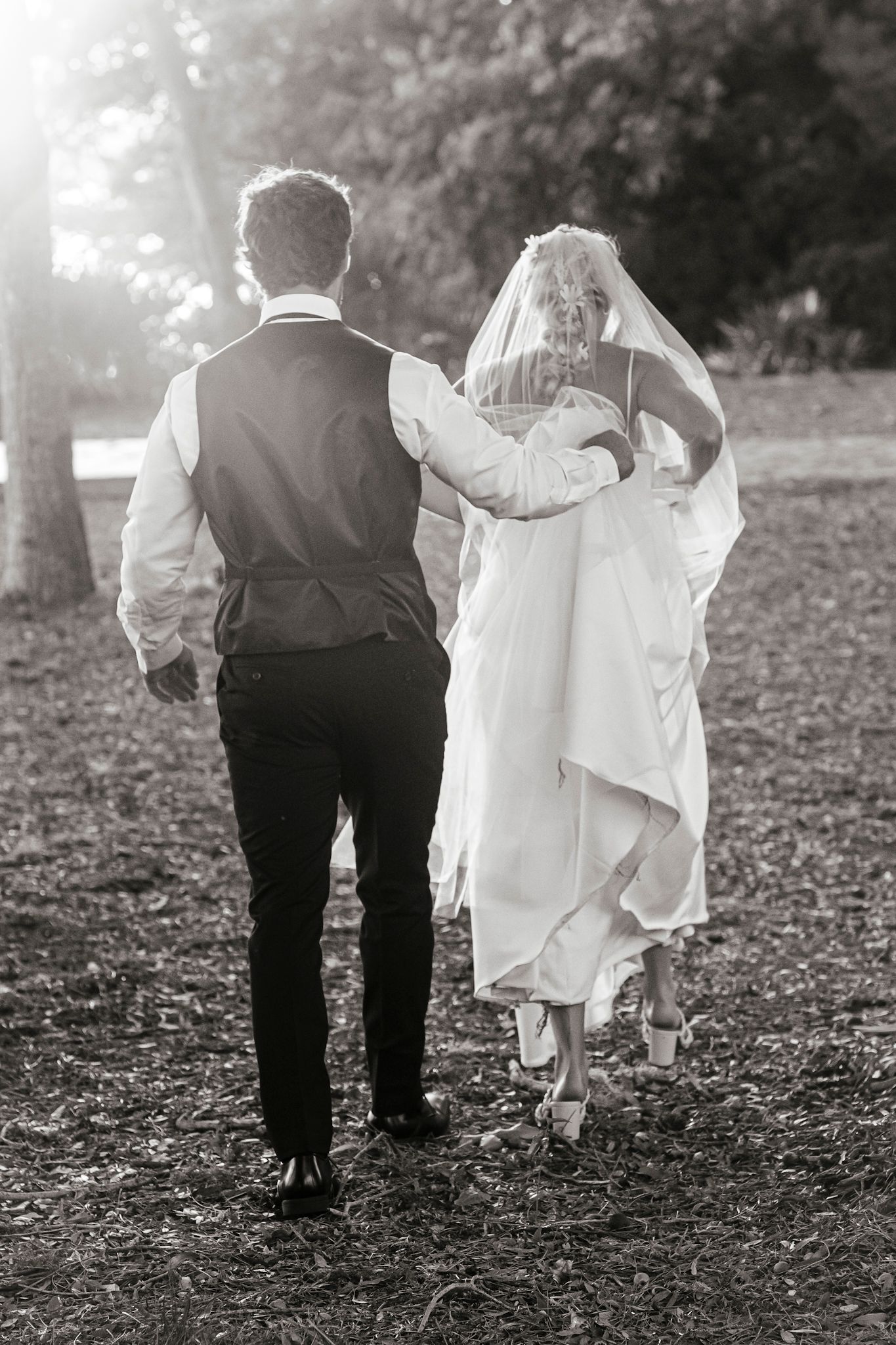 A bride and groom walk away from camera on fallen autumn leaves in black and white vintage style photography.